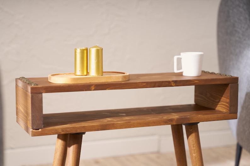 May include: A wooden side table with a shelf and two legs. The table is brown and has a distressed finish. There are three gold candles on a wooden tray on the top shelf.
