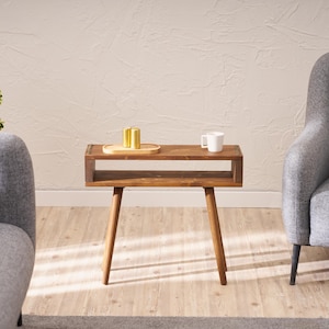 May include: A wooden side table with a shelf and four legs. The table is brown and has a natural wood finish. There is a small tray on the top shelf with three gold-coloured items. A white mug is on the table.