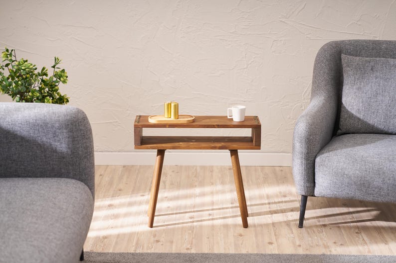 May include: A wooden side table with a shelf and four legs. The table is brown and has a natural wood finish. There is a small tray on the top shelf with three gold-coloured items. A white mug is on the table.