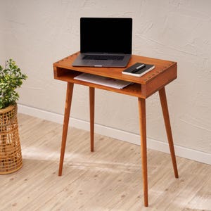 May include: A mid-century modern style wooden desk with a laptop, notebook, and pen on top. The desk has a drawer and four angled legs. A woven basket with a plant is to the left of the desk.