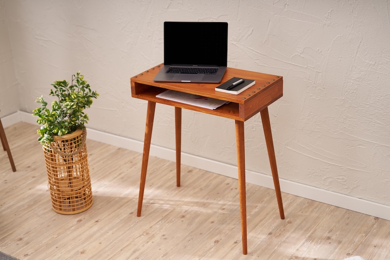 May include: A mid-century modern style wooden desk with a laptop, notebook, and pen on top. The desk has a drawer and four angled legs. A woven basket with a plant is to the left of the desk.