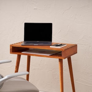 May include: A wooden desk with a laptop, notebook, and pen. The desk has a drawer and is supported by four angled legs. A gray office chair is in the foreground. The desk is made of wood and has a warm, natural finish.
