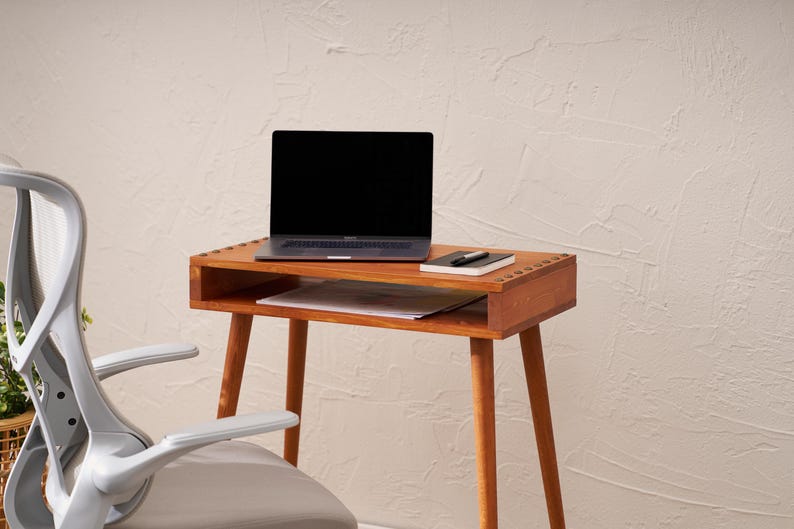 May include: A wooden desk with a laptop, notebook, and pen. The desk has a drawer and is supported by four angled legs. A gray office chair is in the foreground. The desk is made of wood and has a warm, natural finish.