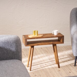 May include: A wooden side table with a shelf and four legs. The table is brown and has a white ceramic mug and three gold metal containers on top.