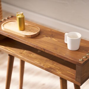 May include: A wooden side table with a shelf and two legs. The table is stained a dark brown colour and has brass accents. A white mug and a small wooden tray with two gold containers are on the table.