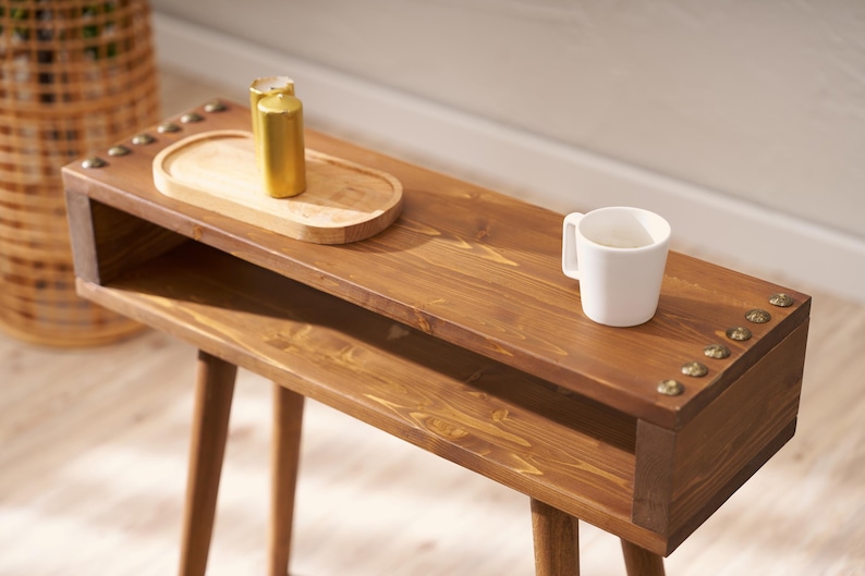 May include: A wooden side table with a shelf and two legs. The table is stained a dark brown colour and has brass accents. A white mug and a small wooden tray with two gold containers are on the table.