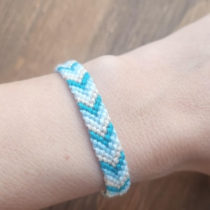 May include: A woven friendship bracelet in light blue, white, and beige. The chevron pattern bracelet is shown on a wrist. This handmade accessory is a stylish piece of jewellery, perfect for everyday wear.