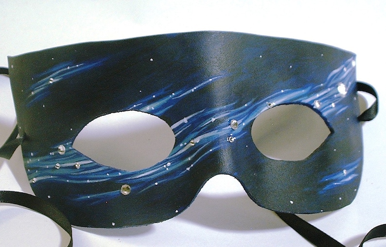 May include: A black leather masquerade mask with blue and white painted designs and silver rhinestones. The mask has two eye holes and a black ribbon tie.