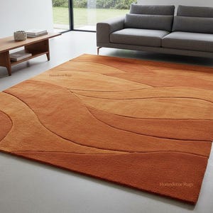 May include: An orange and brown area rug with a wave-like pattern. The rug has a gradient of colors, transitioning from a darker shade of orange at the edges to lighter shades in the center. The text "Homedecor Rugs" is visible.