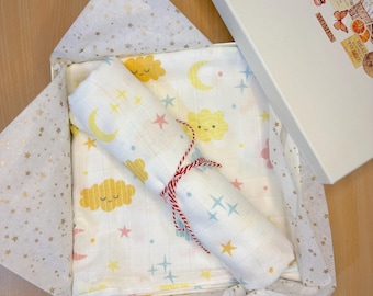 May include: A white muslin swaddle blanket with a yellow, pink and blue cloud and star pattern. The blanket is rolled up and tied with a red and white string. The blanket is in a white gift box with gold star patterned tissue paper.