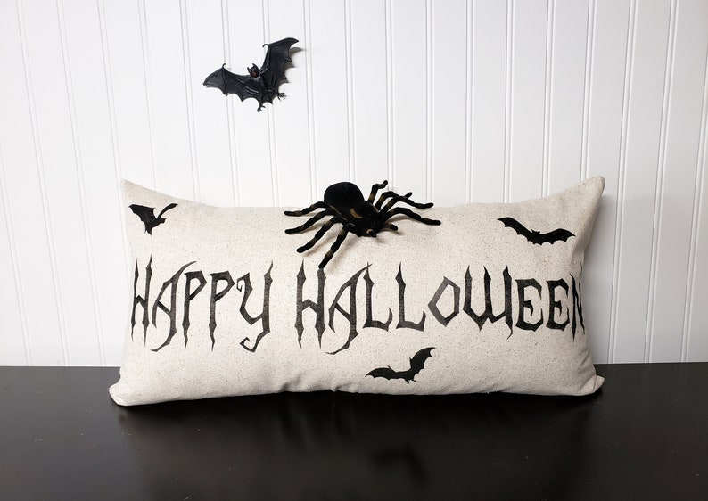 K&ouml;nnte beinhalten: Ein beiges Kissen mit dem Schriftzug "Happy Halloween" in Schwarz. Das Kissen ist mit schwarzen Flederm&auml;usen und einer schwarzen Spinne verziert.