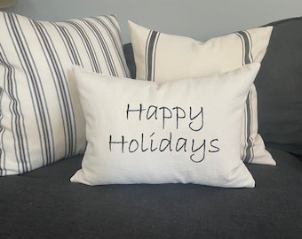 May include: A white throw pillow with black embroidery that reads "Happy Holidays" on a black couch with two striped pillows behind it.