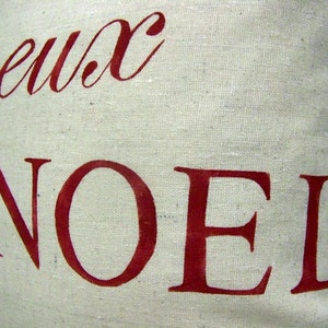K&ouml;nnte beinhalten: Ein wei&szlig;es Kissen mit den Worten "Joyeux Noel" in Rot bedruckt. Die Worte sind in einer kursivschriftlichen Schriftart.