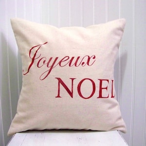 K&ouml;nnte beinhalten: Ein wei&szlig;es Kissen mit der Aufschrift "Joyeux No&euml;l" in roten Buchstaben.