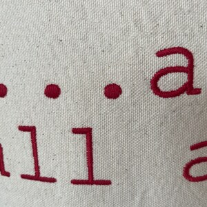 May include: Close-up of a white canvas tote bag with the text "...all are" embroidered in red.