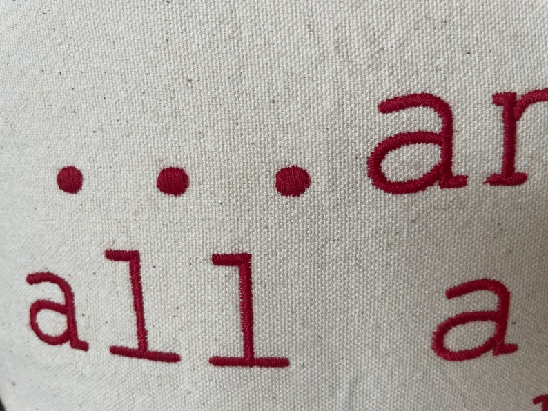 May include: Close-up of a white canvas tote bag with the text "...all are" embroidered in red.