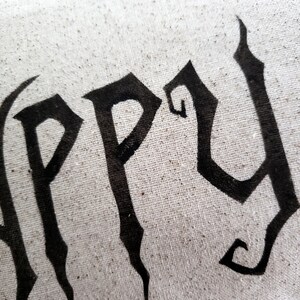 K&ouml;nnte beinhalten: Ein wei&szlig;es Kissen mit dem Wort "Happy" in schwarzen gotischen Buchstaben.