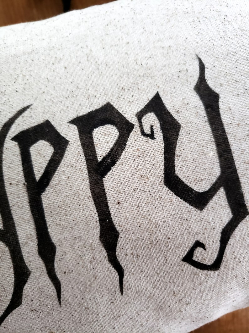 K&ouml;nnte beinhalten: Ein wei&szlig;es Kissen mit dem Wort "Happy" in schwarzen gotischen Buchstaben.