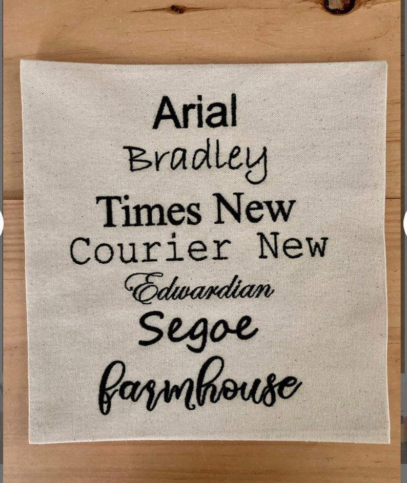May include: A white fabric swatch with different font names embroidered in black thread. The fonts include Arial, Bradley, Times New Roman, Courier New, Edwardian Script ITC, Segoe Script, and Farmhouse.