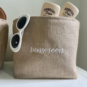 May include: Two brown burlap storage bins with white embroidery. The bin on the right has the word "sunscreen" embroidered on the front and contains a pair of white sunglasses and two bottles of Hawaiian Tropic sunscreen.