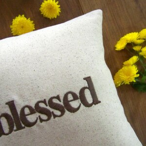 May include: A white pillow with the word "blessed" embroidered in brown on the front. The pillow is surrounded by yellow flowers.