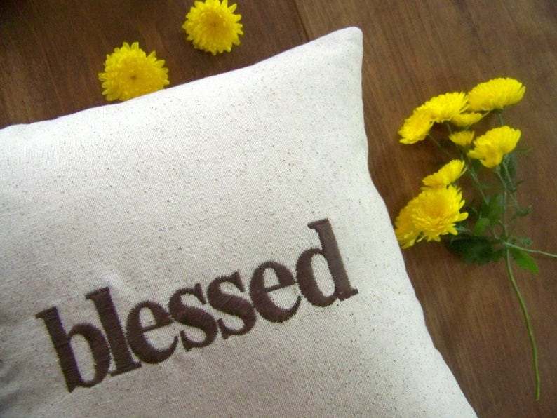 May include: A white pillow with the word "blessed" embroidered in brown on the front. The pillow is surrounded by yellow flowers.