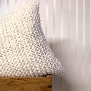 May include: White knitted pillow with a chunky knit texture. The pillow is sitting on a wooden crate.