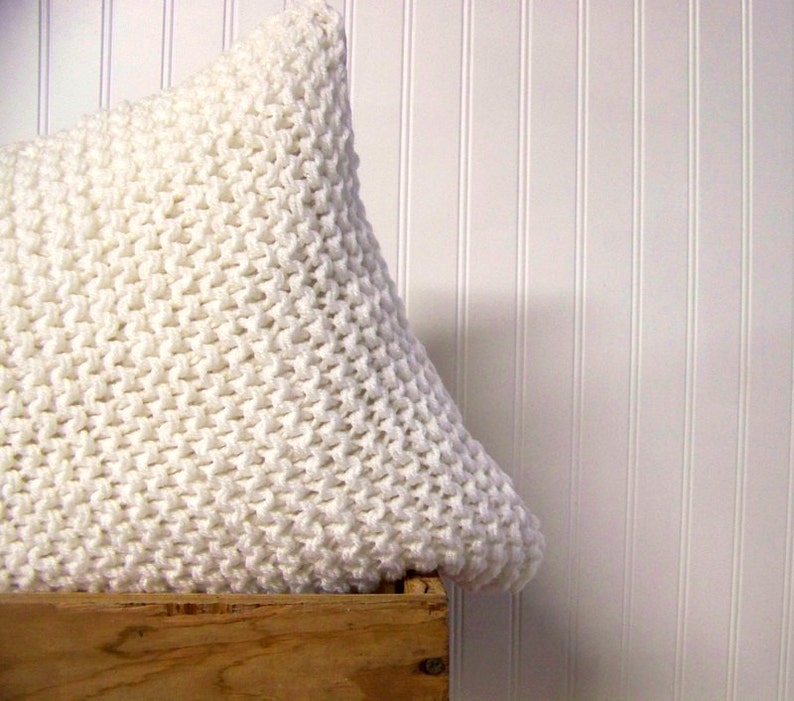 May include: White knitted pillow with a chunky knit texture. The pillow is sitting on a wooden crate.