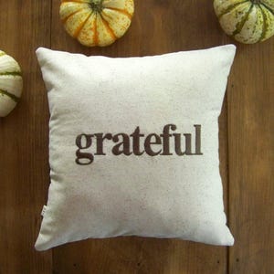 May include: A white throw pillow with the word "grateful" embroidered in brown on a white background. The pillow is surrounded by three small gourds.