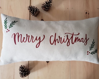 May include: A white rectangular pillow with the text "Merry Christmas" in red cursive lettering. The pillow has green sprigs with red berries on each end.