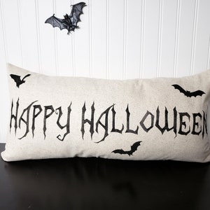 K&ouml;nnte beinhalten: Ein beiger Kissenbezug mit den Worten "Happy Halloween" in schwarzen gotischen Buchstaben. Auf dem Kissen sind vier schwarze Flederm&auml;use.