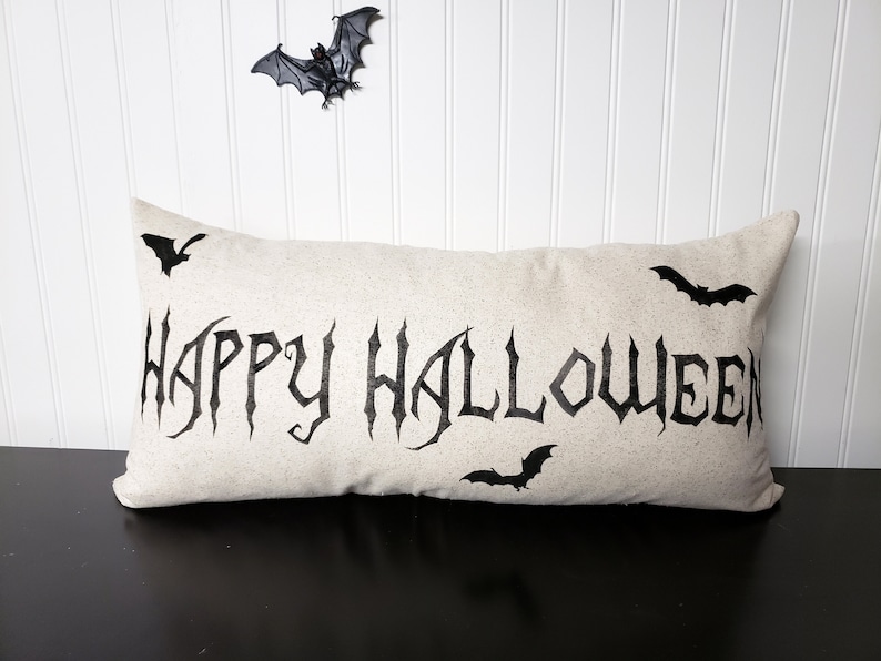 K&ouml;nnte beinhalten: Ein beiger Kissenbezug mit den Worten "Happy Halloween" in schwarzen gotischen Buchstaben. Auf dem Kissen sind vier schwarze Flederm&auml;use.