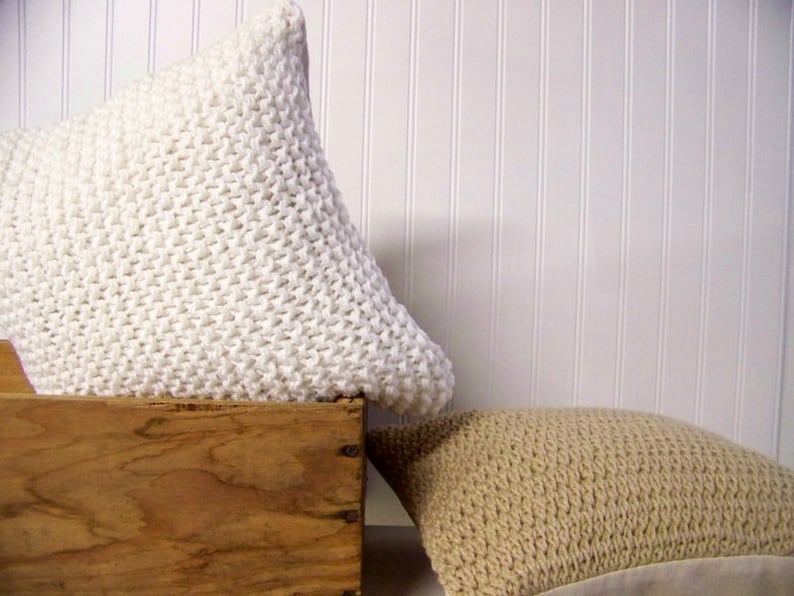 May include: Two knitted throw pillows, one white and one beige, are displayed on a wooden crate. The pillows are made with a chunky knit stitch.