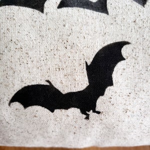 K&ouml;nnte beinhalten: Ein wei&szlig;es Kissen aus Canvas mit einer schwarzen Fledermaus-Silhouette und dem Wort "BOO" in schwarzen Buchstaben.