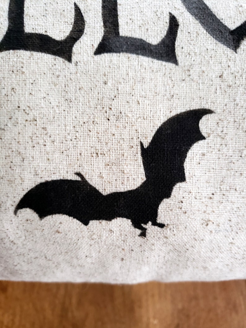 K&ouml;nnte beinhalten: Ein wei&szlig;es Kissen aus Canvas mit einer schwarzen Fledermaus-Silhouette und dem Wort "BOO" in schwarzen Buchstaben.