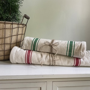 May include: Two linen tea towels, one with red stripes and one with green stripes, are rolled up and tied together with twine. The towels are sitting on a white table.