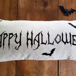 K&ouml;nnte beinhalten: Ein wei&szlig;es Kissen mit den Worten "Happy Halloween" in schwarzer gotischer Schrift. Auf dem Kissen sind vier schwarze Flederm&auml;use gedruckt. Das Kissen liegt auf einer Holzoberfl&auml;che.