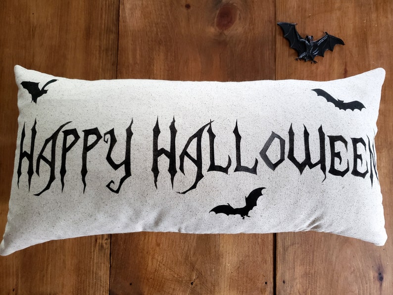 K&ouml;nnte beinhalten: Ein wei&szlig;es Kissen mit den Worten "Happy Halloween" in schwarzer gotischer Schrift. Auf dem Kissen sind vier schwarze Flederm&auml;use gedruckt. Das Kissen liegt auf einer Holzoberfl&auml;che.