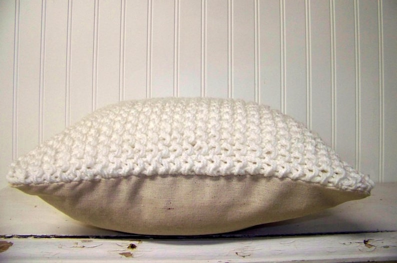 May include: White crocheted pillow with a beige linen backing. The pillow is made with a chunky yarn and has a textured surface.