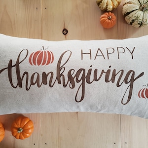 May include: A white pillow with the text "Happy Thanksgiving" printed in brown. The pillow is decorated with two brown and orange pumpkins.