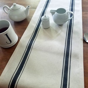 May include: A white table runner with black stripes, a white teacup with the letter J, a white teapot, a white creamer, and a white sugar bowl. The table runner is on a wooden table.