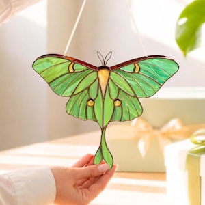 May include: A stained glass luna moth suncatcher. The moth features green wings with yellow accents and a yellow body. The wings are outlined in dark brown. The suncatcher hangs from a white string.