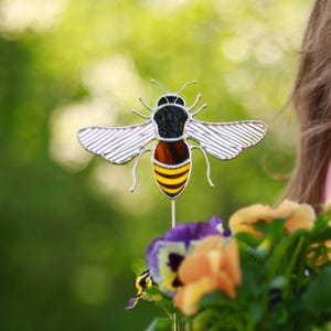 May include: A stained glass bee garden stake with yellow and black stripes, placed in a white pot with a plant with purple and yellow flowers.