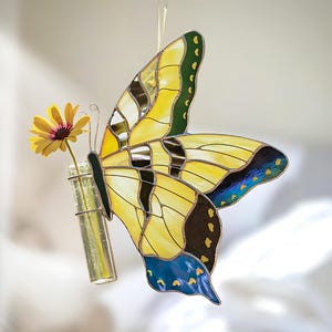 May include: A stained glass butterfly in yellow, black, and blue holds a small glass vial with a yellow flower. The butterfly has delicate wire antennae and is suspended by a clear string, creating a whimsical, decorative piece.