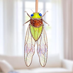 Könnte beinhalten: Ein Glasfenster-Zikaden-Suncatcher mit braunem Körper, grünem Bauch und schillernden Flügeln. Das Insekt hat blaue Augen und einen gelben Kopf. Es hängt an einer braunen Lederschnur, ideal für Fenster- oder Wanddekoration.