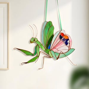Stained glass praying mantis suncatcher hanging on wall with green ribbon, handmade insect glass decor with colorful wings, modern interior wall decoration.