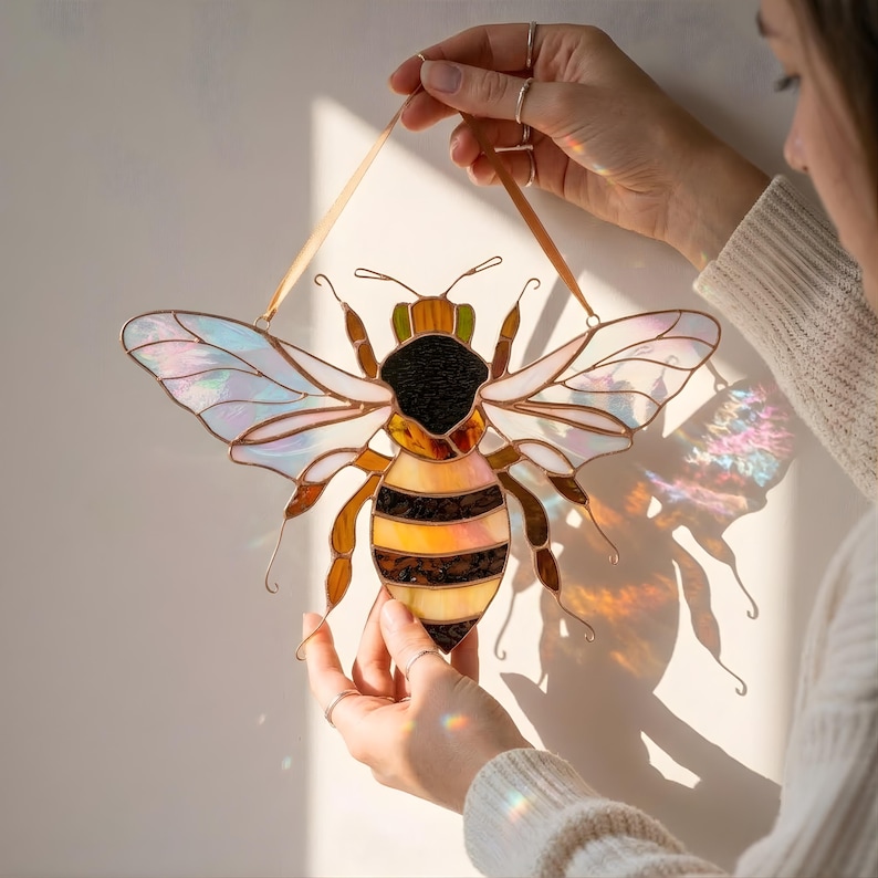 Person hanging handmade stained glass bee suncatcher on wall in sunlight. Iridescent wings cast colorful reflections while amber and brown glass body highlights copper foil craftsmanship.