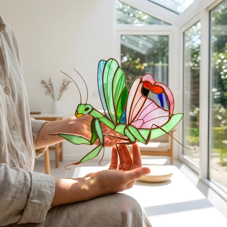 Handmade stained glass praying mantis suncatcher held in hands, showing size and detailed glass wings, unique insect decor made with copper foil technique.