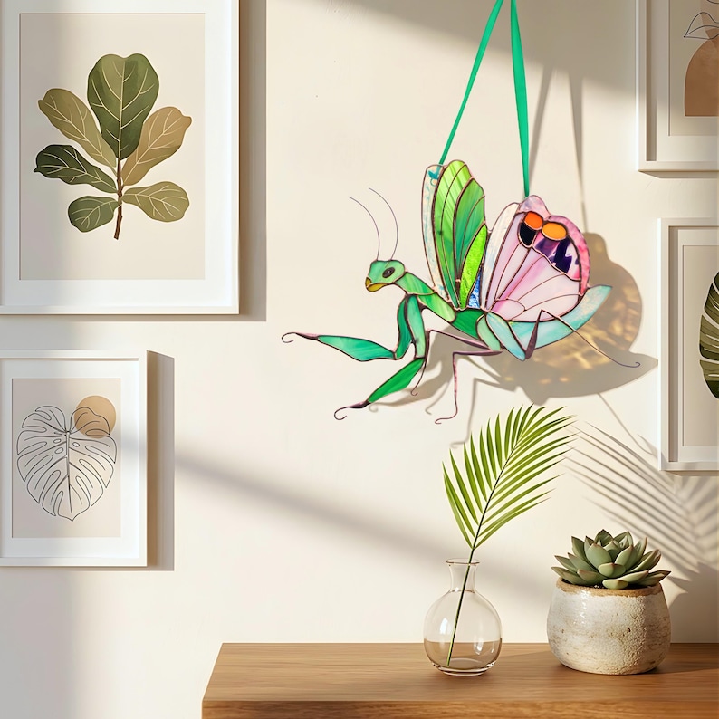 Stained glass praying mantis suncatcher hanging in cozy interior with plants and framed art, colorful insect decor casting soft shadows in sunlight.