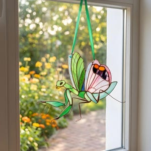 Stained glass praying mantis suncatcher hanging on window, sunlight shining through colorful glass wings, handmade insect decor creating soft light reflections.